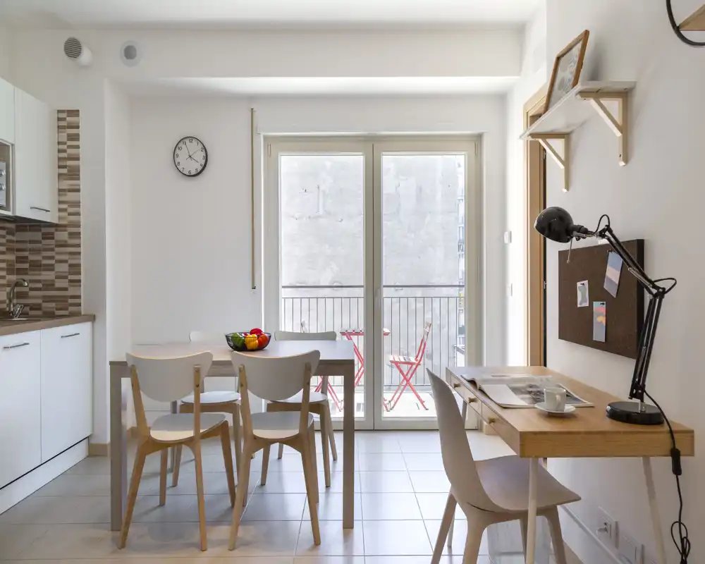 Bright modern dining area adjacent to a compact kitchen, with a simple wooden table, four chairs and large glass doors opening to a small balcony — ideal for casual meals and natural light.