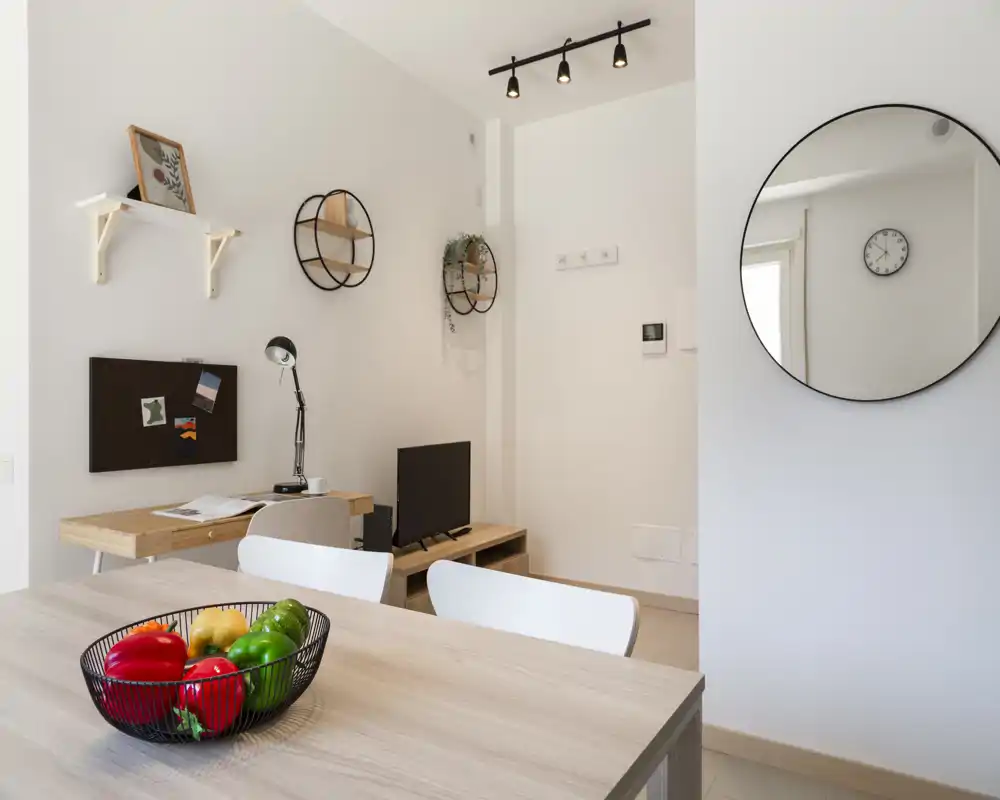 Bright, modern living area with a wooden table in the foreground, decorative wall shelves, a small TV console and a round wall mirror. Clean minimalist styling with natural light and inviting accents like a fruit bowl.