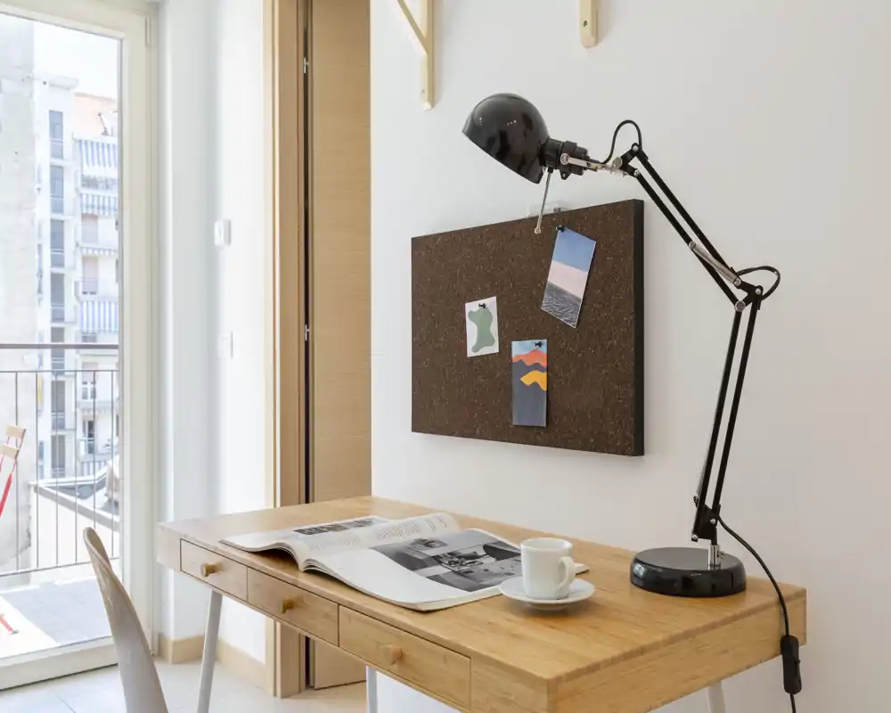 Compact, bright home-office corner with a wooden desk, adjustable black desk lamp, pinboard and a balcony visible through sliding glass doors.
