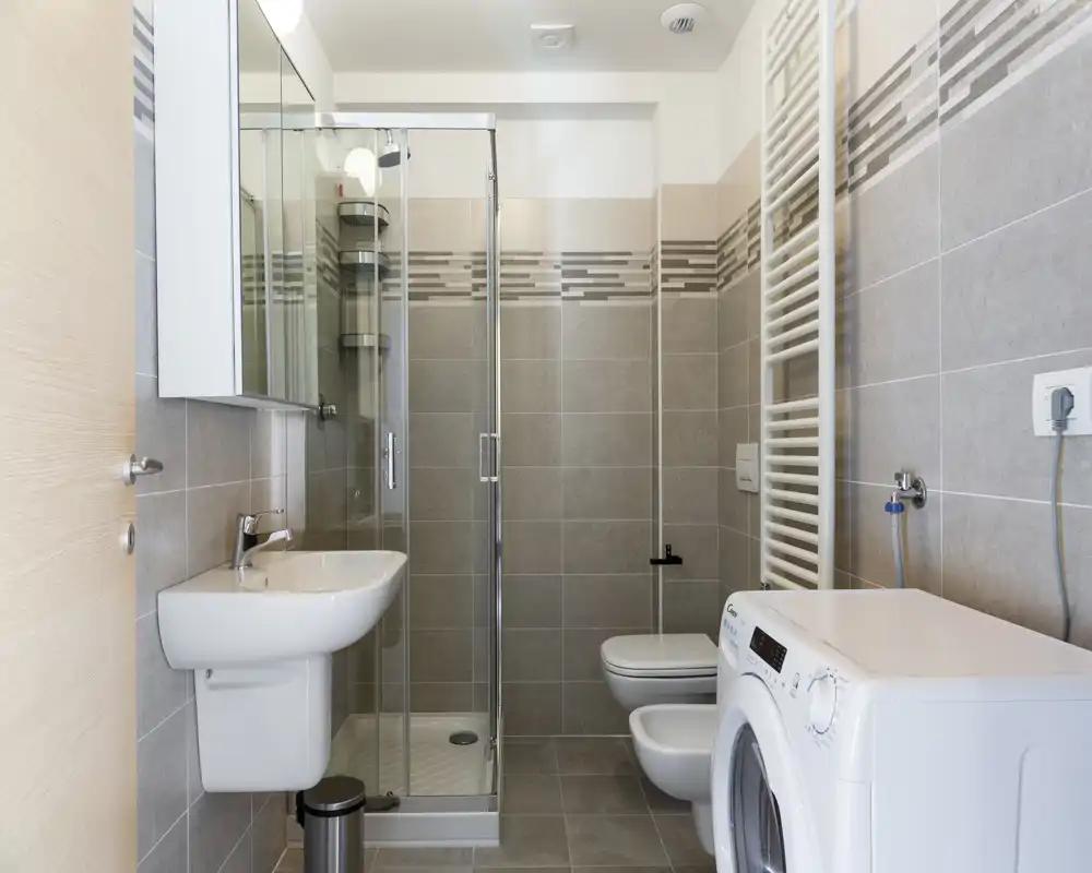 Modern compact bathroom with a glass shower enclosure, wall-mounted sink, bidet and toilet, and a washing machine neatly placed to the side. Neutral grey tiles and clean lines give a fresh, functional feel.