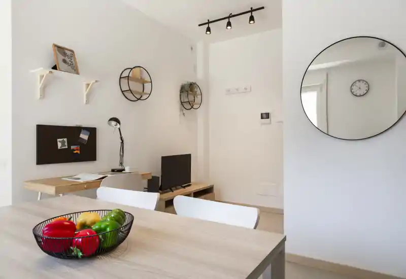 Bright, modern living area with a wooden table in the foreground, decorative wall shelves, a small TV console and a round wall mirror. Clean minimalist styling with natural light and inviting accents like a fruit bowl.