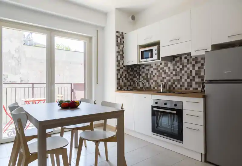 Cocina moderna y luminosa con mesa comedor, cuatro sillas y salpicadero de azulejos; grandes puertas correderas se abren a una pequeña terraza con luz natural.