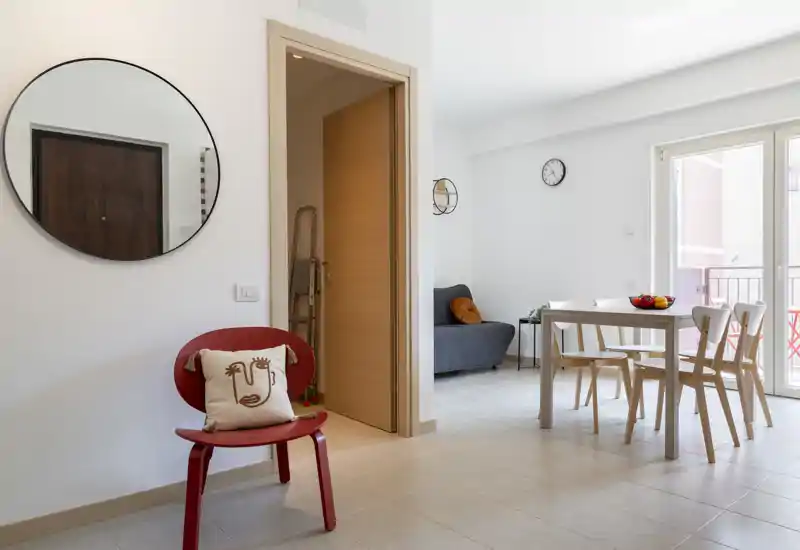 Bright, modern living/dining area with a small dining table, chairs and a seating nook by the balcony. Clean lines, neutral tones and decorative accents (mirror, cushion) create a welcoming open-plan space.