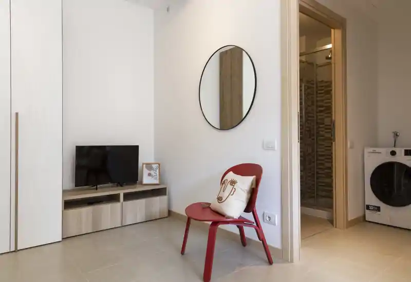 Simple, modern living area with a red accent chair, wall mirror, low TV cabinet and neutral tile flooring — bright and tidy space ideal for short stays.