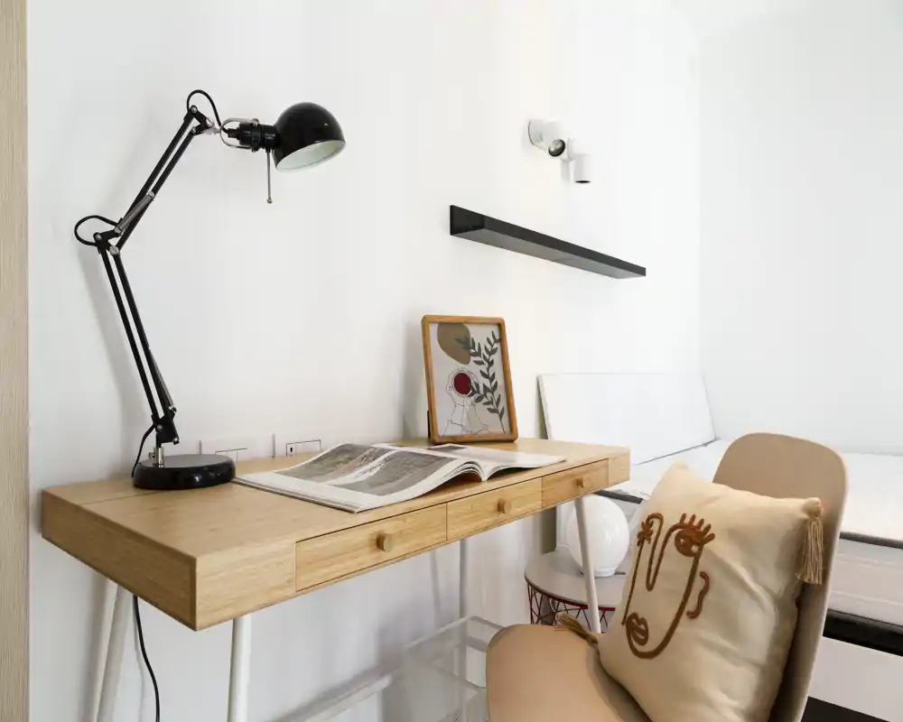 Coin chambre lumineux et minimaliste avec bureau en bois, lampe de lecture et coussin décoratif ; un lit partiellement visible à l’arrière-plan indique un espace nuit compact et chaleureux.