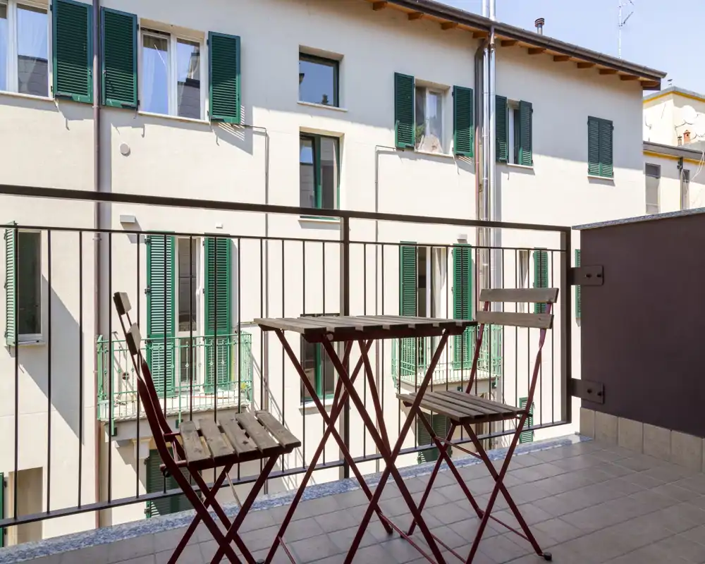Petit balcon ensoleillé avec une table bistro en bois et deux chaises pliantes, sol carrelé et garde-corps métallique donnant sur les volets du bâtiment d'en face.