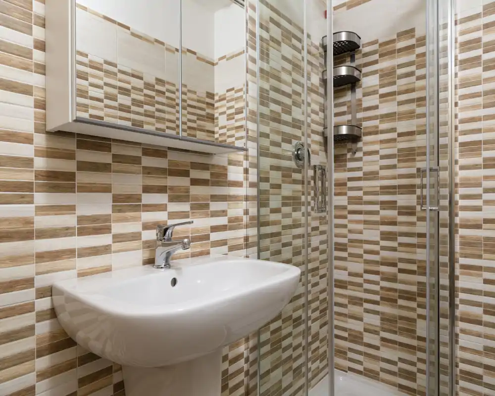 Salle de bain compacte et moderne avec lavabo sur colonne, armoire miroir et douche à portes vitrées, carrelage beige et blanc à motif.