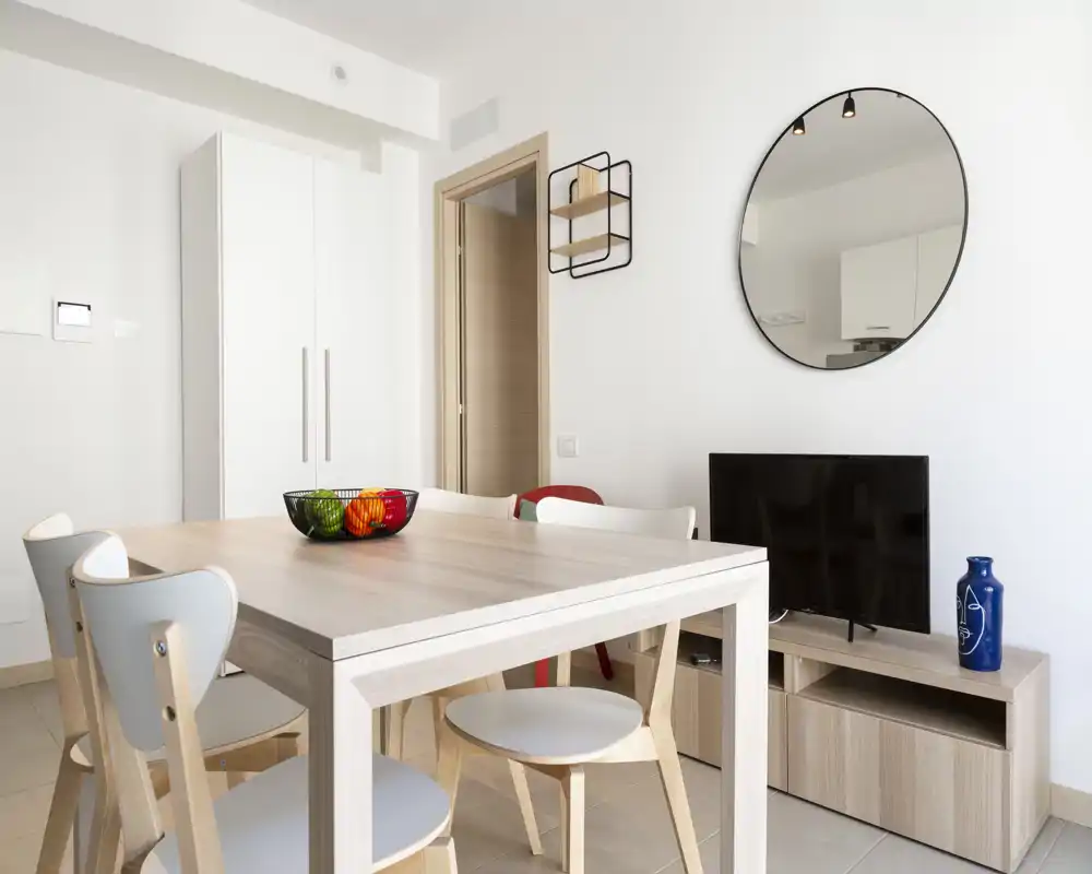 Coin repas moderne et lumineux avec une table en bois clair, chaises assorties et un bol de fruits décoratif ; décoration minimaliste et miroir mural qui agrandit l'espace.