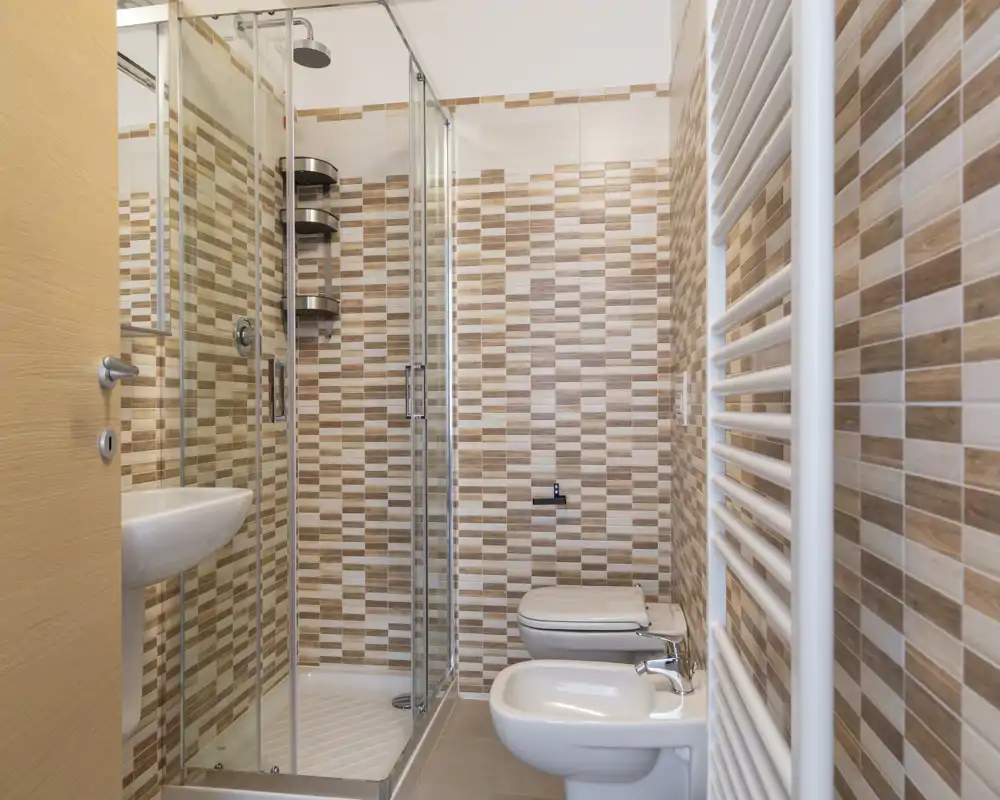 Salle de bain moderne carrelée avec douche vitrée, lavabo mural, bidet et WC ; carreaux mosaïque beige chaud et sèche-serviettes.