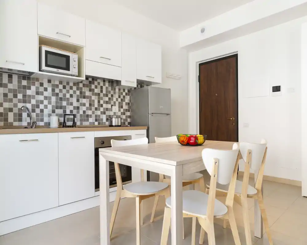 Cuisine moderne ouverte avec table en bois clair et quatre chaises, placards blancs, crédence carrelée et réfrigérateur inox — lumineuse, soignée et prête à être utilisée.
