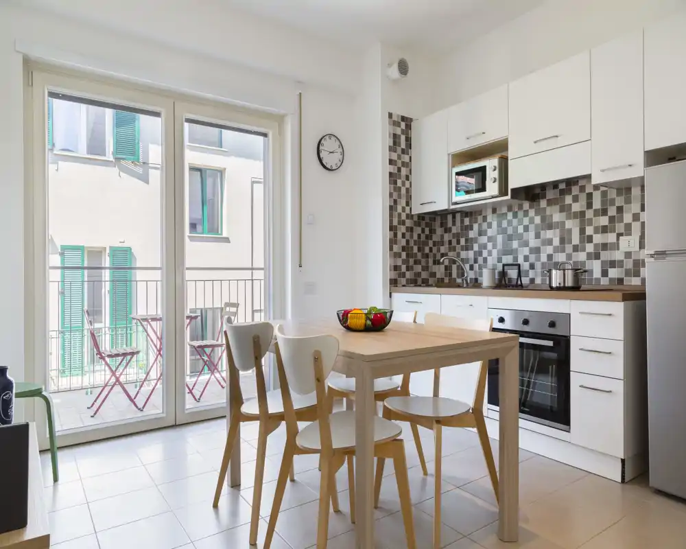 Cuisine moderne et lumineuse avec table en bois, quatre chaises et un plan de travail équipé d'un four, micro-ondes et réfrigérateur ; grandes portes vitrées donnant sur un petit balcon apportant beaucoup de lumière naturelle.