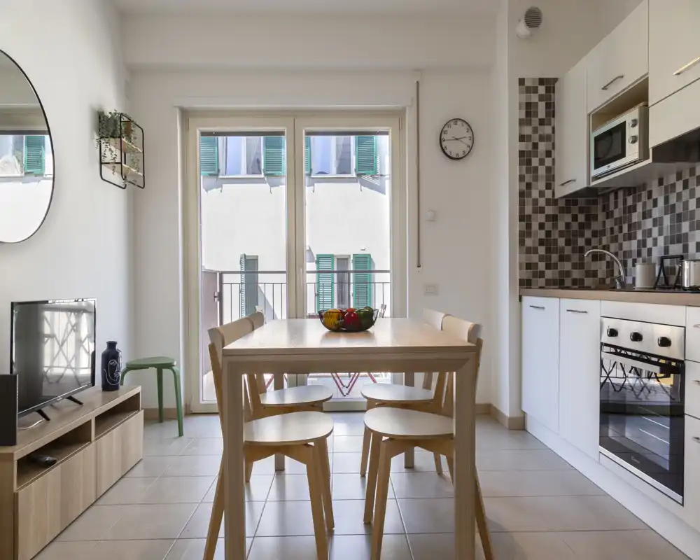 Cuisine moderne et lumineuse en open-space avec table en bois, quatre chaises et électroménagers intégrés ; grande fenêtre apportant de la lumière naturelle et une ambiance cosy et minimaliste.