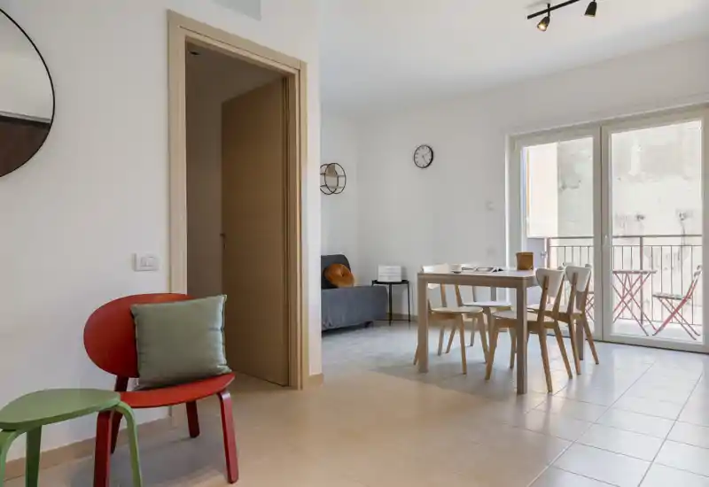 Espacio salón/comedor luminoso y moderno con un sofá pequeño, mesa para cuatro y grandes puertas correderas hacia el balcón. Mobiliario minimalista de estilo escandinavo y detalles cálidos que crean un ambiente acogedor.