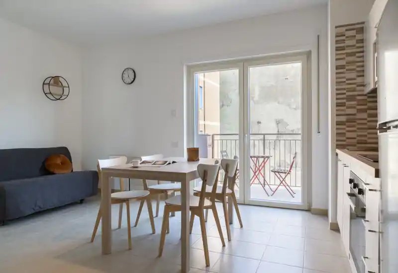 Zona de estar luminosa y moderna con mesa de comedor para cuatro, sofá compacto y grandes puertas de cristal que dan a un pequeño balcón. Colores neutros y decoración minimalista que crean un ambiente tranquilo y acogedor.