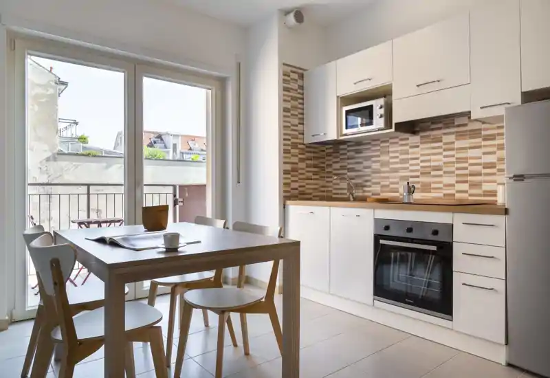 Cocina moderna y luminosa con mesa de madera, cuatro sillas, horno y microondas integrados y grandes puertas de cristal que dan a un pequeño balcón — ideal para comidas con mucha luz.