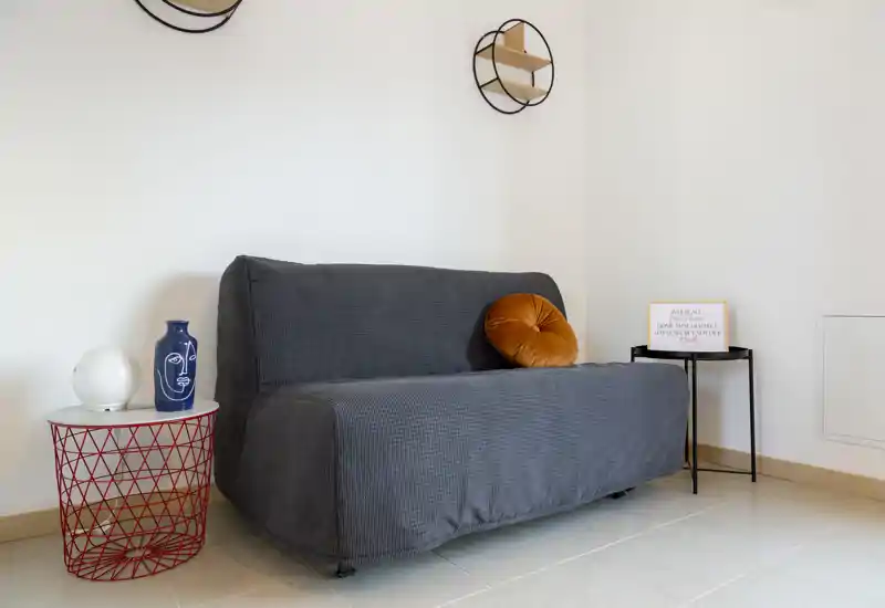 Área de salón compacta y moderna con un sofá gris tapizado, cojines decorativos y mesitas auxiliares; estilo luminoso y minimalista, ideal para un estudio acogedor.