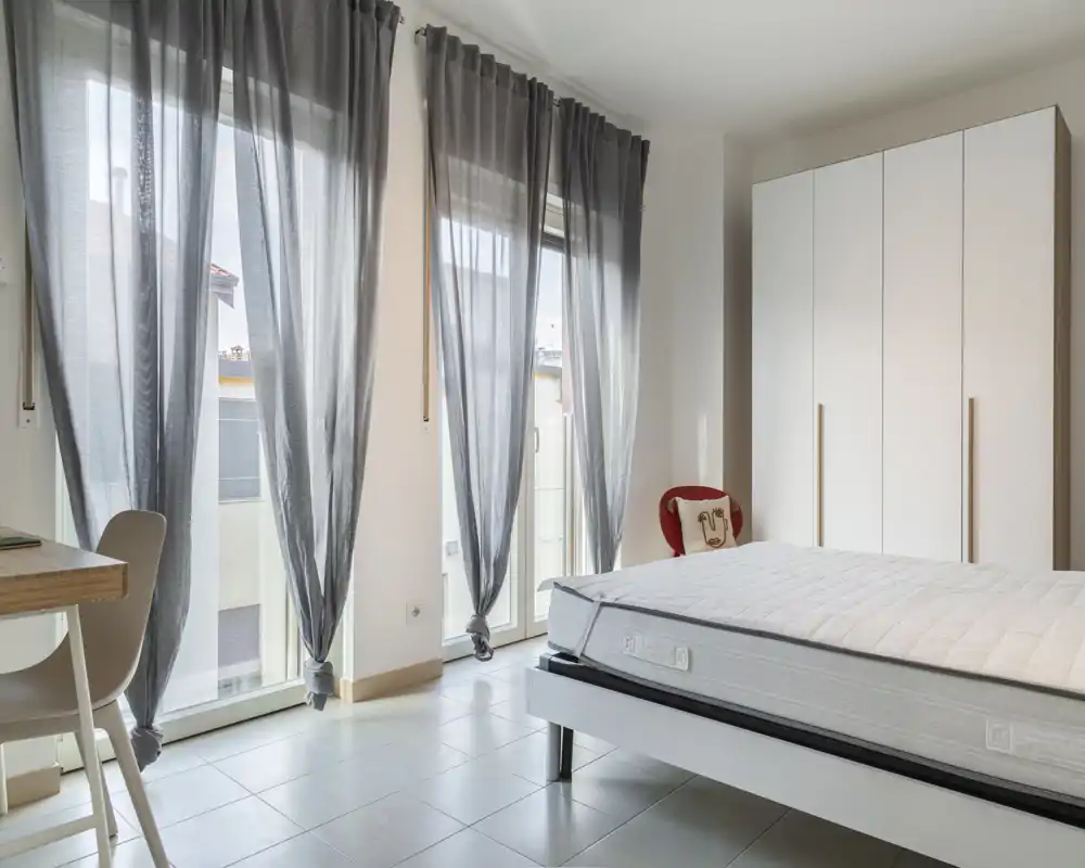 Dormitorio moderno y luminoso con grandes ventanas de suelo a techo con cortinas grises translúcidas, un armario blanco y un colchón sobre una cama baja — ambiente aireado, minimalista y bien iluminado.