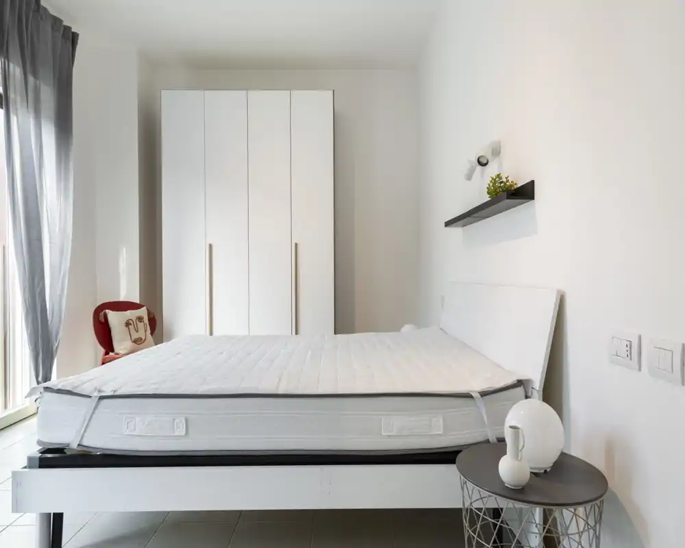 Dormitorio moderno y luminoso con una gran cama blanca y armario; decoración minimalista, luz natural desde la ventana izquierda, mesita y estante con detalles sutiles.