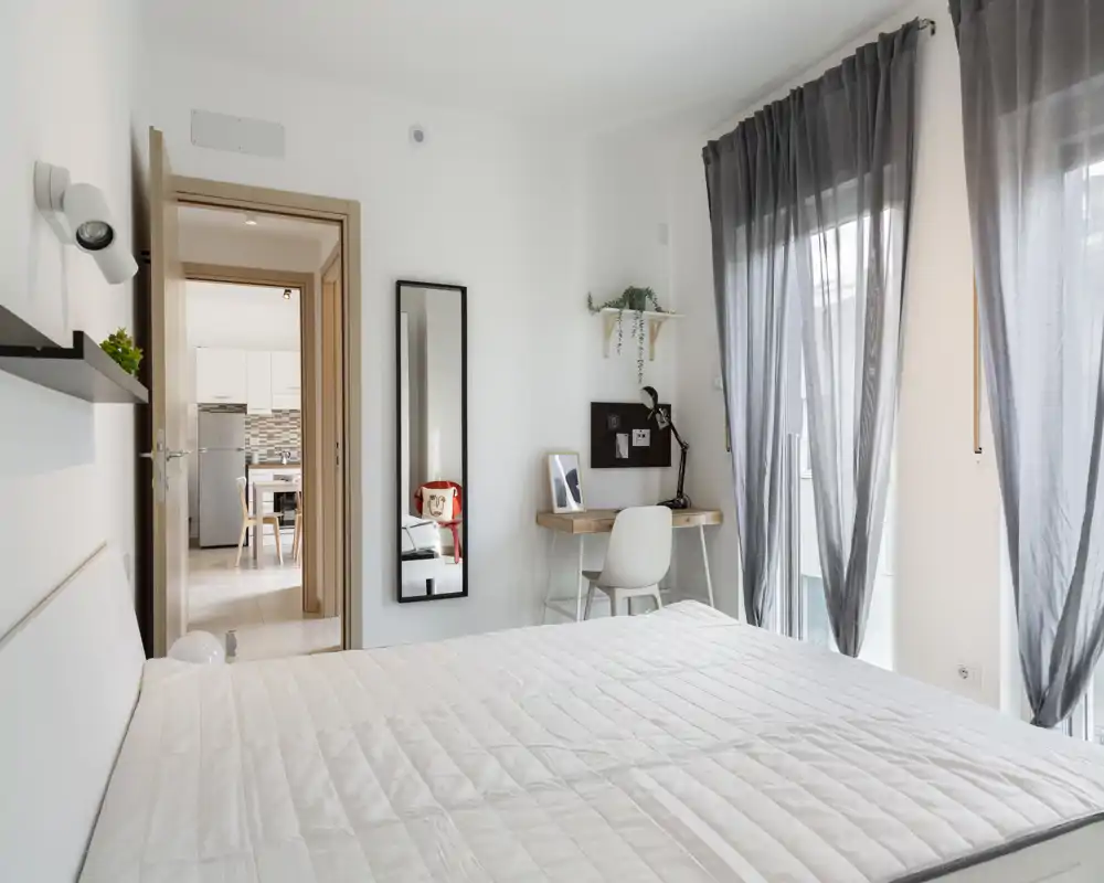 Dormitorio moderno y luminoso con una gran cama en primer plano, un pequeño espacio de trabajo con escritorio y silla junto a la ventana y cortinas translúcidas que dejan pasar la luz natural.