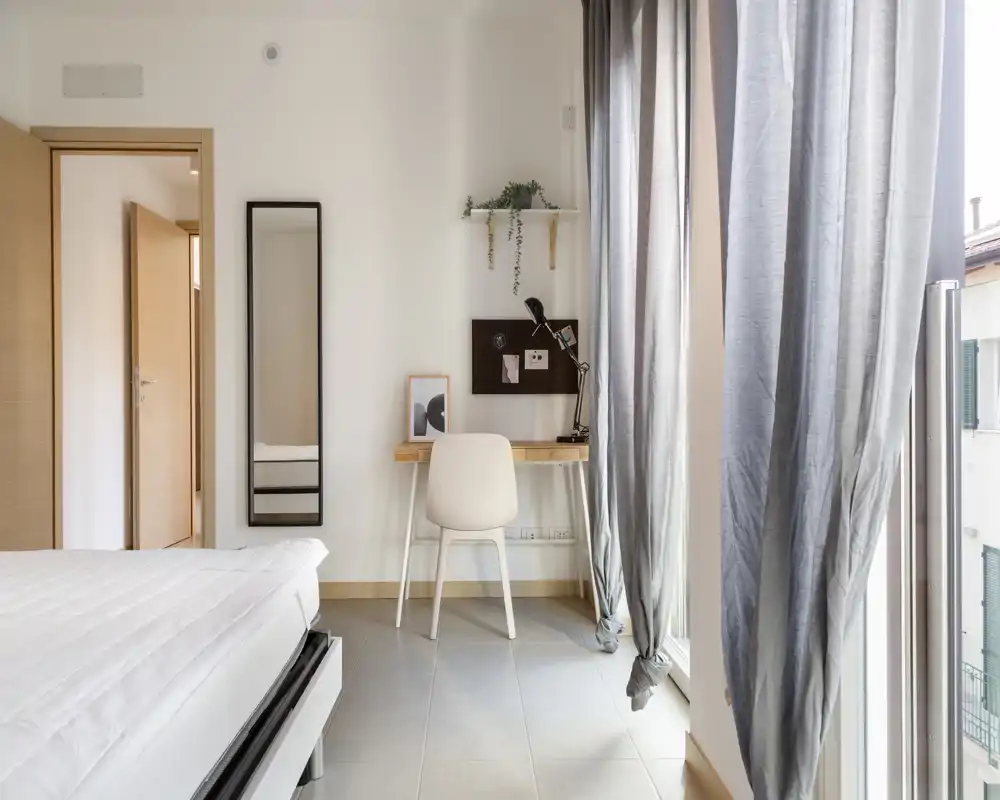 Dormitorio luminoso y minimalista con colchón visible y una pequeña zona de trabajo junto a la ventana. Las cortinas grandes permiten la entrada de luz natural; ambiente moderno y aireado.