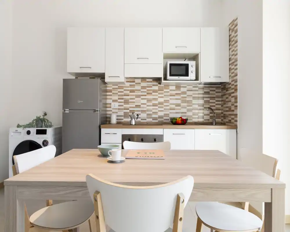 Cocina compacta moderna y luminosa con muebles blancos, salpicadero con patrón y mesa de madera en primer plano; incluye nevera, microondas y lavadora — ideal para pisos pequeños.