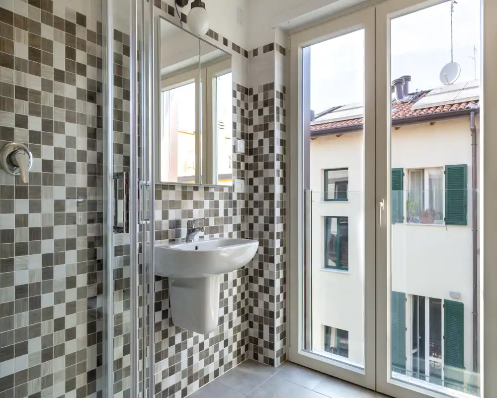 Baño moderno y luminoso con paredes de mosaico, lavabo suspendido, mampara de ducha de cristal y una gran ventana que aporta luz natural.