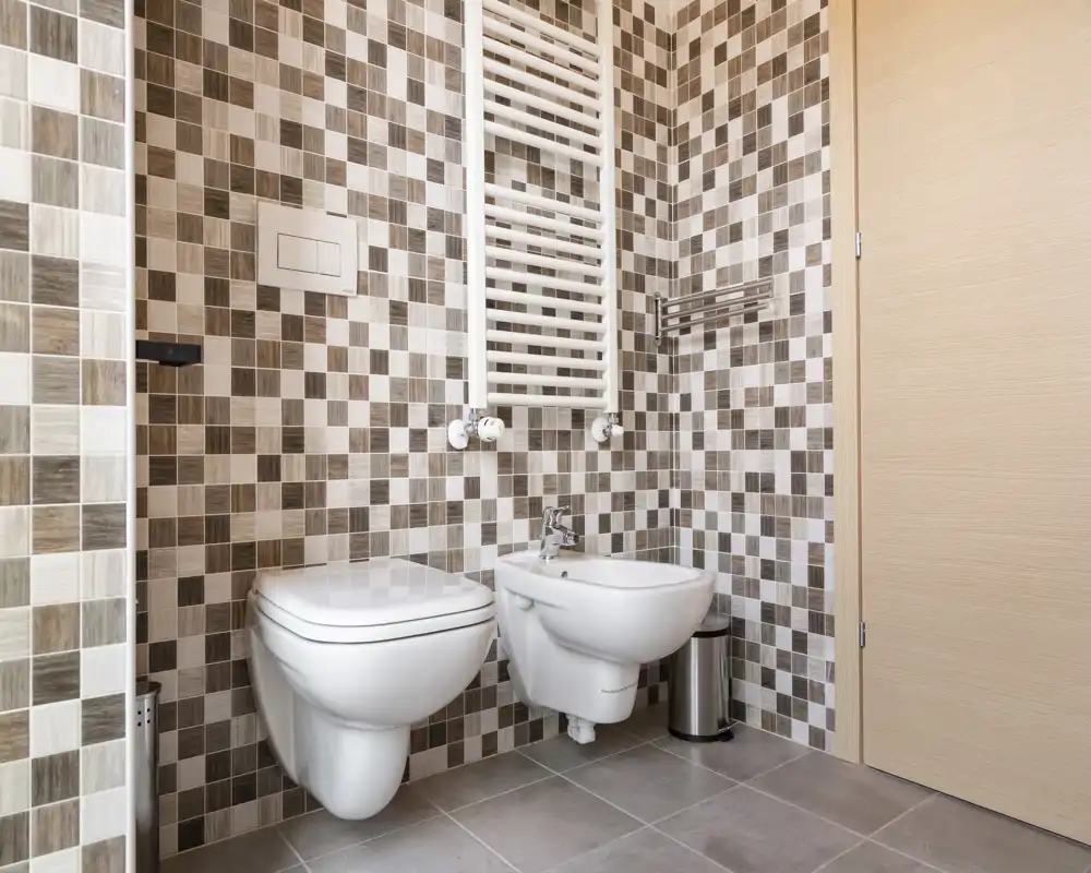 Baño moderno con azulejos en mosaico, inodoro suspendido y bidé, grifería cromada y toallero calefactado que crea un aspecto limpio y contemporáneo.
