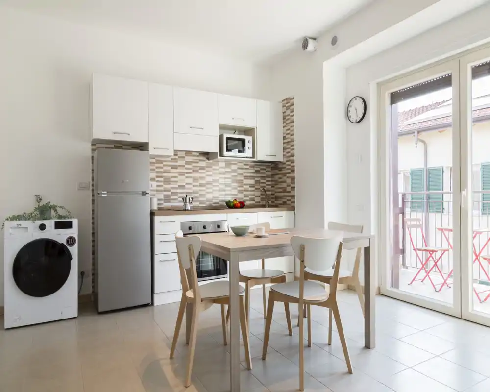 Cocina moderna y luminosa en espacio abierto con armarios blancos, salpicadero de azulejos, nevera, lavadora y una pequeña mesa de comedor junto a una gran puerta corredera hacia el balcón.