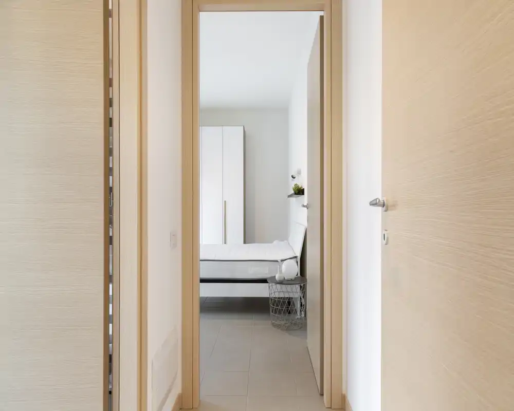 Dormitorio luminoso y minimalista visto desde el marco de la puerta, con cama blanca, armario y mesita sencilla: líneas modernas y luz natural.