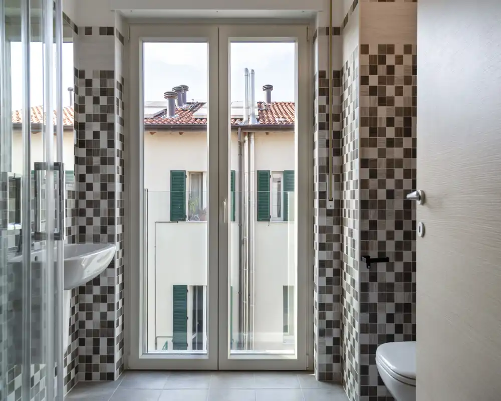 Baño moderno y luminoso con paredes de mosaico, ducha de vidrio, lavabo mural e inodoro, y una gran ventana que aporta luz natural.