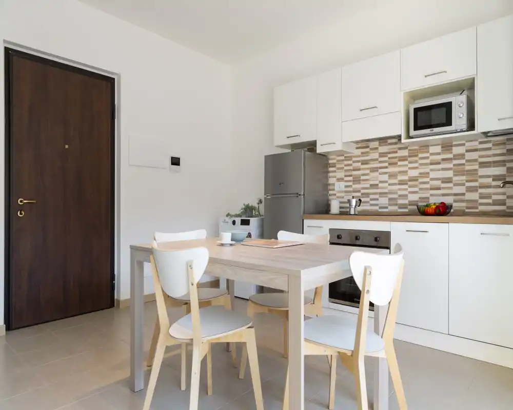 Cocina moderna y luminosa con armarios blancos, salpicadero de azulejos, frigorífico inoxidable y mesa con cuatro sillas — ideal para un apartamento compacto.
