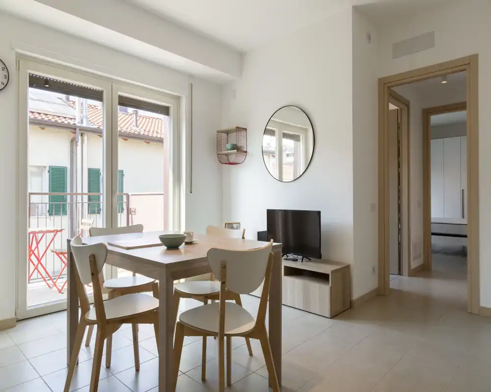 Zona de estar/comedor luminosa y minimalista con mesa de madera y cuatro sillas, luz natural desde las grandes puertas correderas que dan a un pequeño balcón. Decoración neutra, espejo en la pared y mueble de TV compacto, ambiente moderno y tranquilo ideal para alquileres temporales.