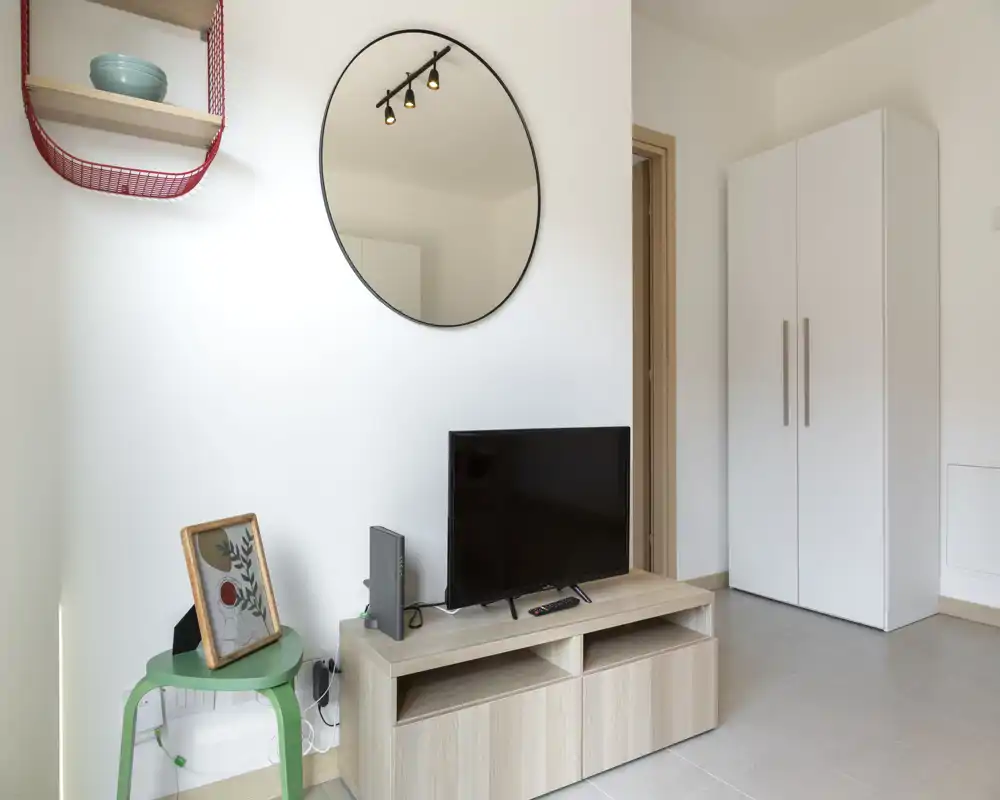 Espacio de estar luminoso y minimalista con mueble de TV compacto, espejo decorativo y armario — un rincón limpio y moderno ideal para relajarse o ver la televisión.