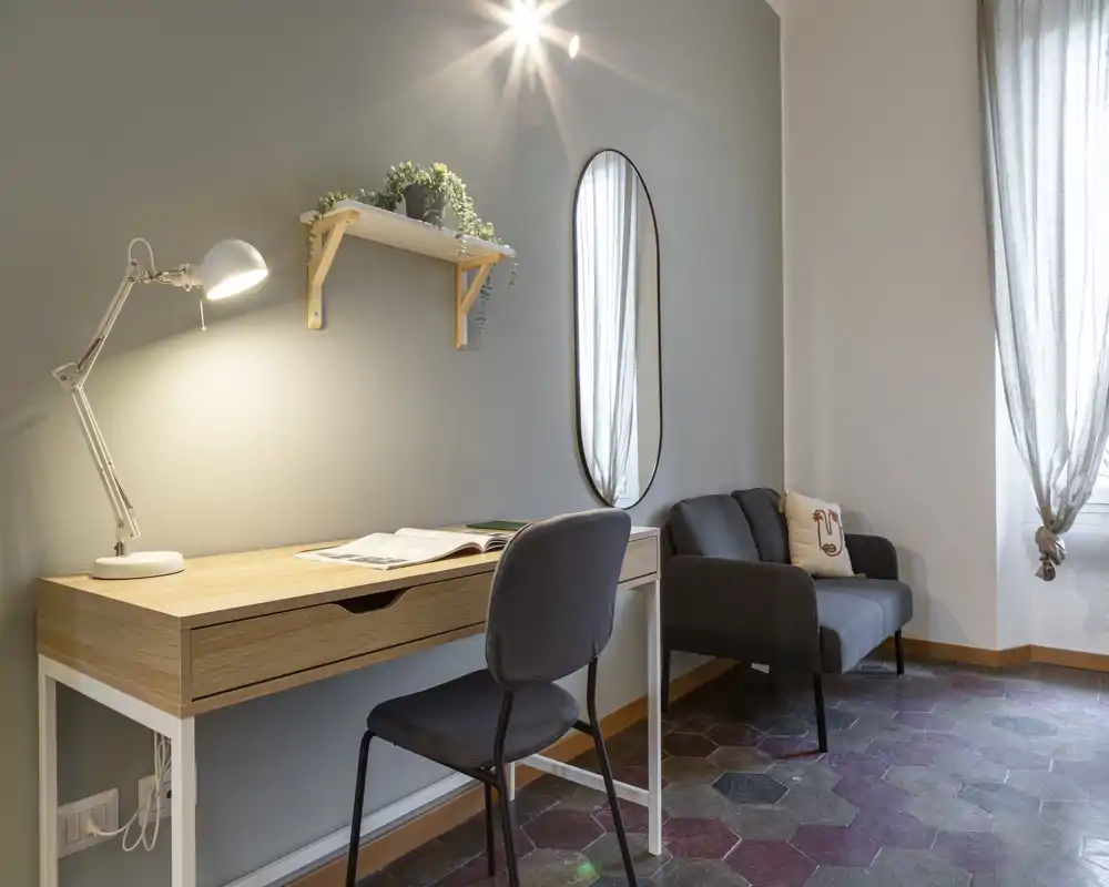 Zona de estar compacta y elegante con un escritorio de madera, lámpara de trabajo y un pequeño sofá tapizado junto a la ventana — espacio luminoso para trabajar y relajarse.