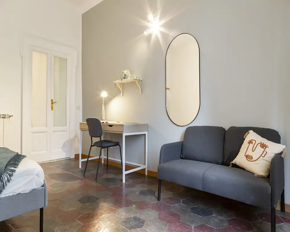 Dormitorio luminoso y con estilo, con cama parcialmente visible, escritorio compacto con lámpara y un sofá pequeño; iluminación cálida y espejo elegante que crean un ambiente acogedor.