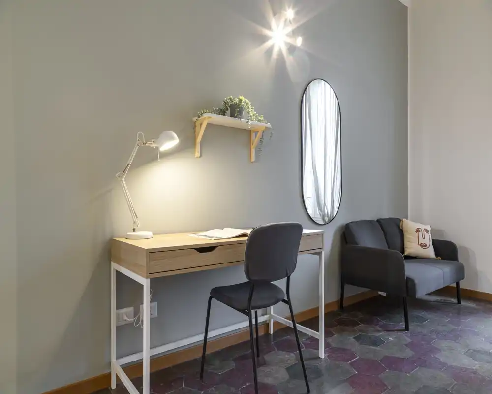 Acogedor rincón de sala moderno y luminoso con un sofá compacto y un escritorio de madera iluminado por una lámpara de mesa — ideal para teletrabajo o relajarse en un piso pequeño.