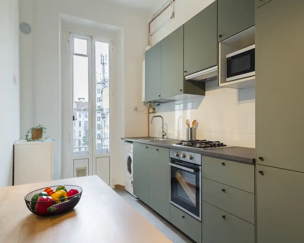 Cocina moderna y luminosa con armarios verdes mate, horno empotrado y vitro de gas, fregadero y lavadora bajo la encimera; una mesa de madera con un cuenco de frutas aporta calidez y luz natural procedente de las altas ventanas.