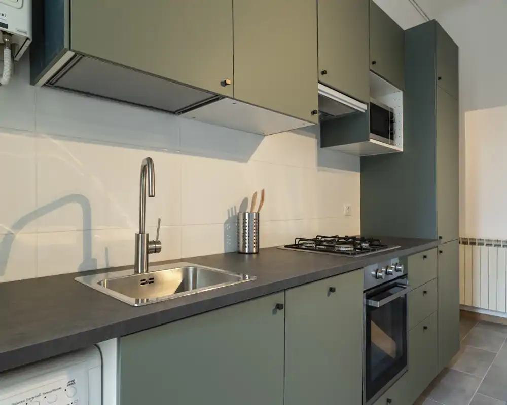 Cocina moderna y compacta con muebles verdes mate, horno integrado y placa de gas, fregadero de acero inoxidable y encimera minimalista — luminosa, limpia y práctica para el día a día.