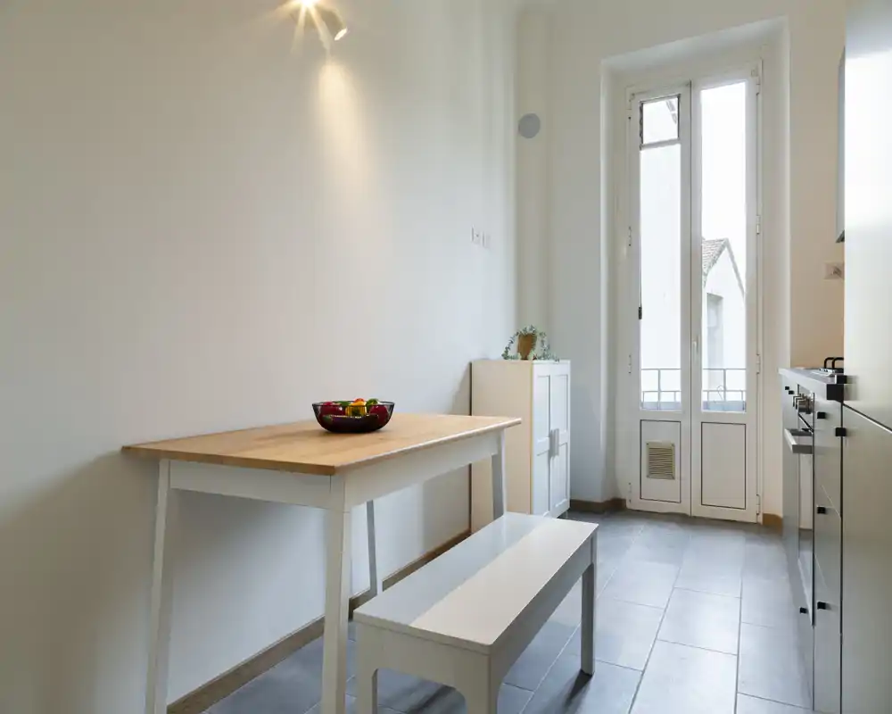 Área de comedor minimalista y luminosa con mesa de madera, banco y un cuenco de frutas; amplia ventana/puerta proporciona mucha luz natural.