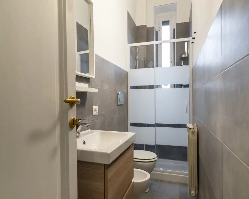 Baño moderno y compacto con lavabo sobre mueble, inodoro, bidé y ducha con mampara de vidrio; baldosas grises y una ventana alta y estrecha que aporta luz natural.