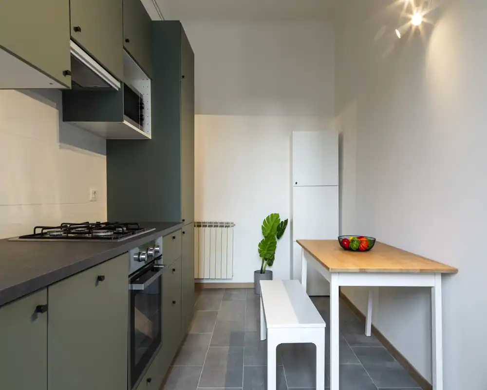 Cocina compacta y moderna con armarios verdes mate, horno integrado y placa de gas, mesa de madera con banco y planta decorativa — un espacio ordenado y funcional para cocinar y comer informalmente.