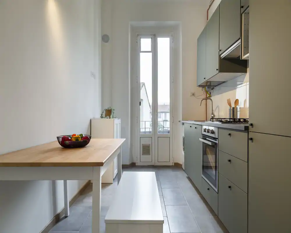 Cocina de diseño en galería con armarios verde apagado, horno integrado y placa de gas, mesa con superficie de madera; luz natural intensa por las puertas altas.