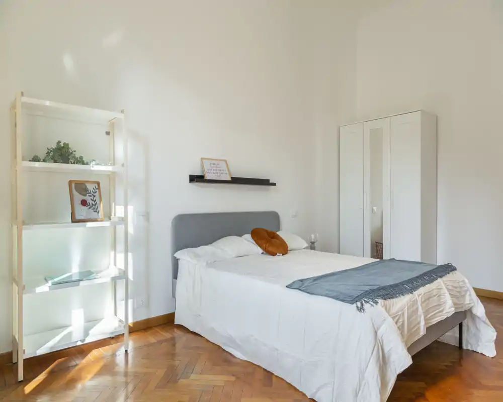 Dormitorio luminoso y minimalista con cama doble, cabecero gris, armario y estantería. La luz natural realza el suelo de madera y los textiles simples y acogedores.