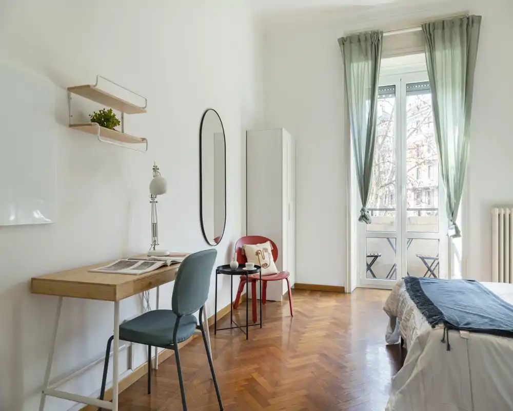 Dormitorio luminoso y ventilado con cama bien hecha, suelo de parquet y un pequeño escritorio junto a la ventana — ideal para trabajar y relajarse con balcón y luz natural.