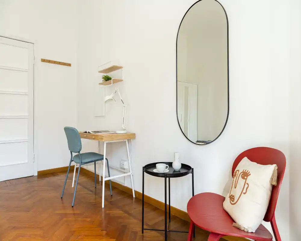 Rincón luminoso y minimalista con una silla roja de acento, una pequeña mesa negra y un escritorio junto a la pared: ideal para relajarse o trabajar ligeramente.