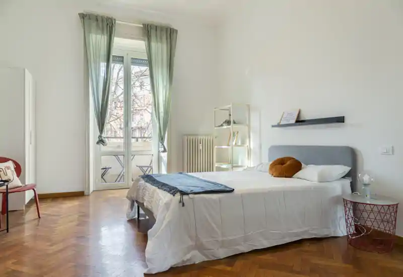 Dormitorio luminoso y amplio con cama doble, cabecero gris, cojín redondo decorativo y una gran ventana que da a un pequeño balcón. Suelo de parquet claro, estanterías minimalistas y detalles cuidados crean un espacio acogedor ideal para anuncios de alquiler.