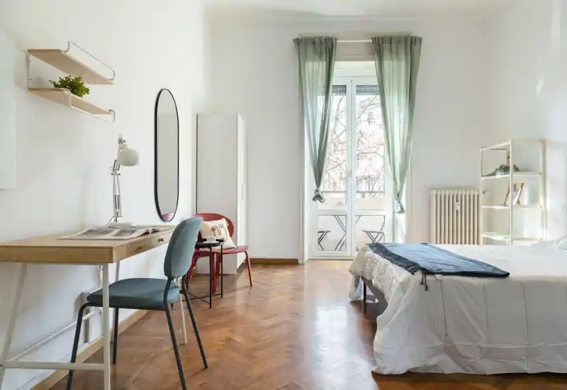 Dormitorio luminoso y ordenado con una cama doble vestida con ropa blanca, un escritorio junto a la pared y una puerta-balconcito que llena la estancia de luz natural.