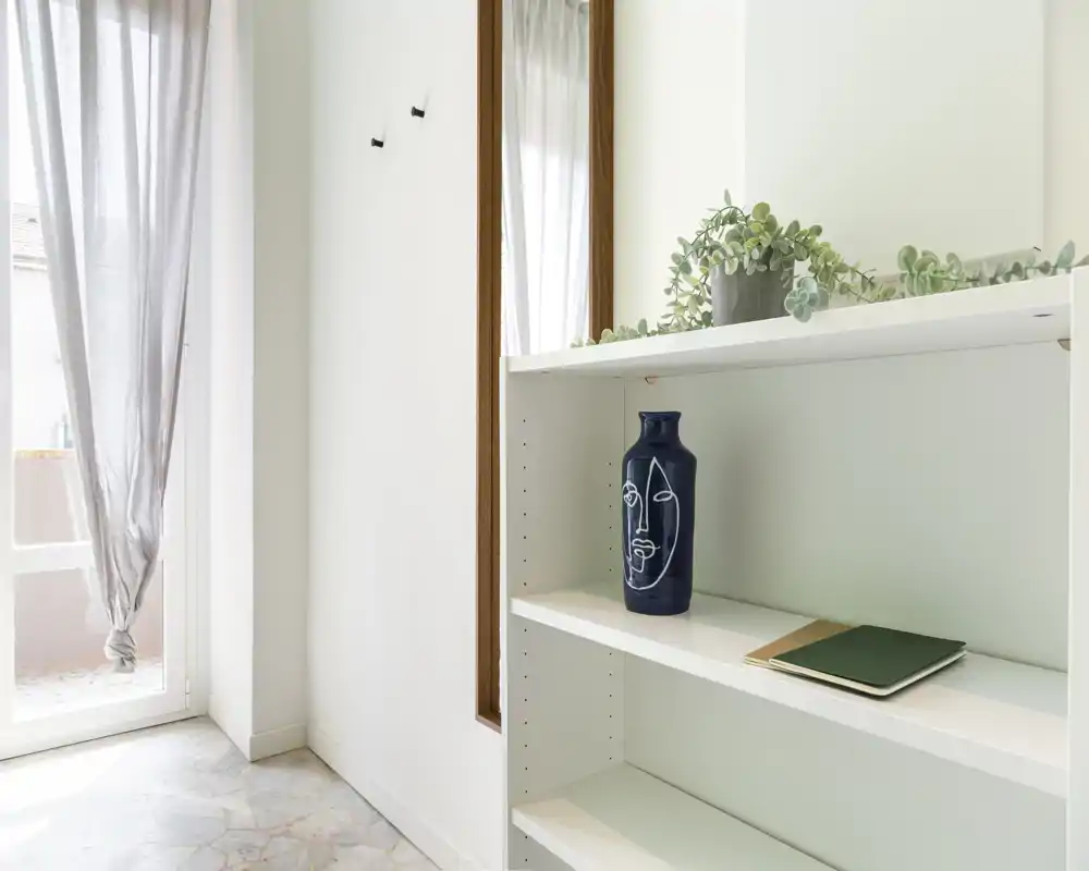 Ingresso luminoso e minimal con una scaffalatura, un vaso decorativo e un grande specchio accanto a una porta con tenda che lascia entrare luce naturale.