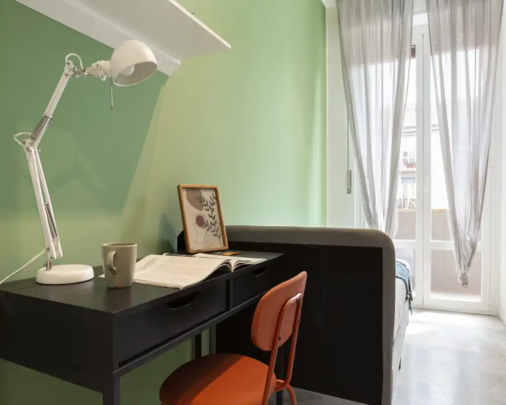 Angolo camera compatto con scrivania e letto parzialmente visibile accanto alla porta-balcone luminosa. La postazione ha una scrivania nera, lampada bianca regolabile e una sedia arancione che si abbinano alla parete verde e alle tende leggere.