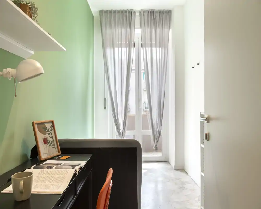 Camera compatta/angolo notte con un piccolo divanetto/letto contenitore visibile, scrivania con sedia, luce naturale dal balcone e arredo moderno e minimale.